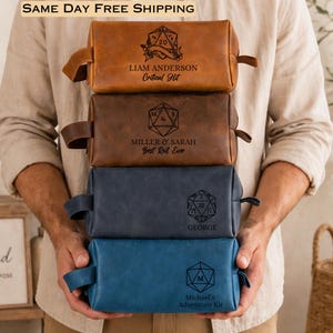 May include: A stack of four rectangular leather toiletry bags in tan, brown, navy, and light blue. Each bag features a side handle and a laser-engraved design of a twenty-sided die with personalized names and phrases like Critical Hit.