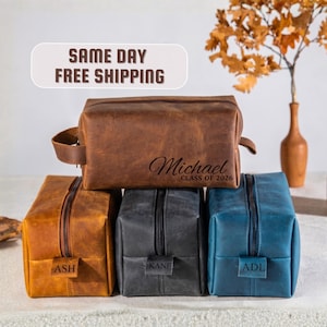 May include: Leather toiletry bags in brown, gold, gray, and blue. The brown bag is personalized with the name "Michael" and "Class of 2026." The other bags are labeled "ASH," "KANE," and "ADL." Each bag has a zipper closure.
