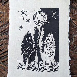 Peut inclure: Gravure sur linoléum en noir et blanc de deux loups debout côte à côte sous une pleine lune. Le loup de gauche est noir et le loup de droite est blanc. L'arrière-plan est un ciel noir et blanc avec des étoiles.