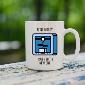 Könnte beinhalten: Weißer Keramikbecher mit einer blau-schwarzen Grafik eines 3D-Druckers. Der Becher trägt den schwarzen Text "DONT WORRY I CAN PRINT A NEW ONE". Der Becher hat einen großen Henkel und steht vor einem verschwommenen grünen Hintergrund.