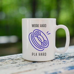 Könnte beinhalten: Weiße Keramiktasse mit einer blauen Grafik einer Spule und den Worten "WORK HARD" und "PLA HARD" in Schwarz. Die Tasse hat einen großen Henkel und steht vor einem verschwommenen grün-weißen Hintergrund.