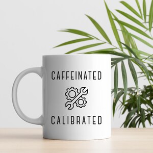 Könnte beinhalten: Weiße Keramiktasse mit den schwarzen Worten "CAFFEINATED CALIBRATED" und einer Zahnrad- und Schraubenschlüssel-Grafik. Die Tasse hat einen gebogenen Henkel und steht vor einem neutralen Hintergrund mit grünem Laub.