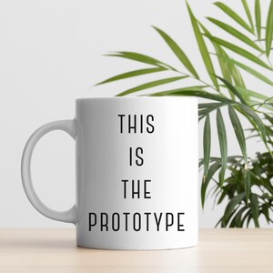Puede incluir: Taza de cerámica blanca con el texto "THIS IS THE PROTOTYPE" en negro, centrado en la parte delantera. La taza tiene un asa curva y se apoya sobre una superficie de madera de color claro. En el fondo se ven hojas verdes de plantas.