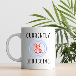 以下が含まれることがあります： 「CURRENTLY DEBUGGING」の黒い文字と、青い円の中に赤い虫のアイコンが描かれた白いセラミックマグカップ。 マグカップは、明るい茶色の表面に置かれ、背景には緑の葉の植物があります。
