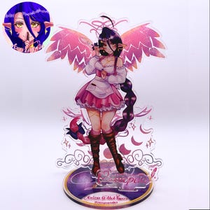 May include: An acrylic standee featuring an anime-style character with long dark hair, pointed ears, and pink wings. The character wears a white blouse, pink skirt, and brown boots. The base is circular with the text Cassiopeia and Cobra's Gilded Tavern.