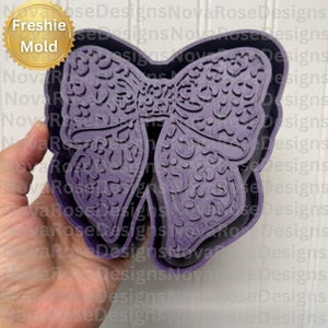 May include: A purple bow-shaped mold with intricate floral patterns. The mold has a layered design and is held in a hand. The text "Freshie Mold" is in a gold circle in the upper left corner.