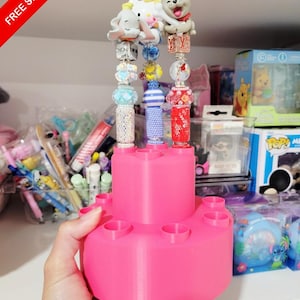 May include: Three decorative pens with character toppers featuring Dumbo, Hello Kitty, and a dog holding a bone. The pens are adorned with various colorful beads and sit in a bright pink, tiered, circular plastic pen holder with multiple slots.