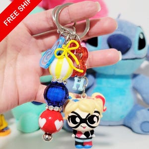 May include: A colorful keychain featuring a Harley Quinn figurine, a blue bead, a red bead with white hearts, and other decorative elements. The keychain has a silver ring and a small letter "H" charm. The background includes a blue stuffed animal.