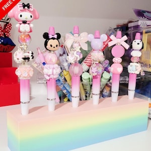 May include: Six pens with ombre pink and white barrels, each topped with decorative beads and character figures including My Melody, Minnie Mouse, and Snoopy, displayed in a rectangular pastel rainbow-colored wooden stand.