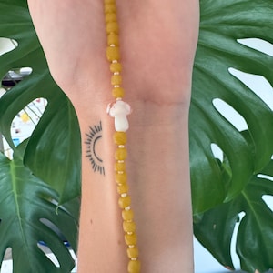 May include: A yellow beaded necklace with a white mushroom charm. The necklace is worn on a person's wrist.