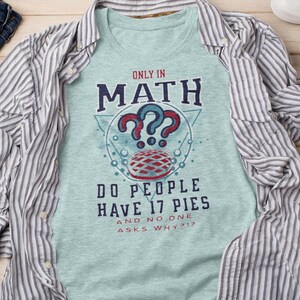 Op de afbeelding: Een lichtblauw T-shirt met een grafisch ontwerp met de tekst "Only in Math do people have 17 pies and no one asks why?!?" De grafiek bevat een rode en witte taart, vraagtekens en een geometrisch ontwerp.