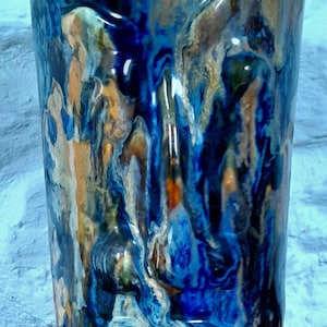 May include: A tall, cylindrical ceramic vase with an abstract design in blue, orange and white. The vase has a textured surface and a glossy finish.
