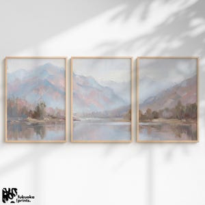 Pode incluir: Conjunto de três impressões de arte de paisagens emolduradas. Cada impressão apresenta uma pintura suave e impressionista de uma cordilheira refletida num lago. A paleta de cores inclui azuis, rosas e castanhos suaves, com uma moldura de madeira clara. As impressões são de fukuoka prints.