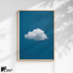 Puede incluir: Impresión enmarcada que presenta una nube blanca y esponjosa contra un cielo azul vibrante. La obra de arte está encerrada en un marco de madera de color claro, creando una estética minimalista. Una serena pieza de arte mural.