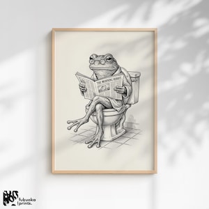 May include: Black and white framed print of a frog sitting on a toilet, reading a newspaper titled "The Morning Ribbit". The frog is wearing a robe. The print is in a light-colored wooden frame.