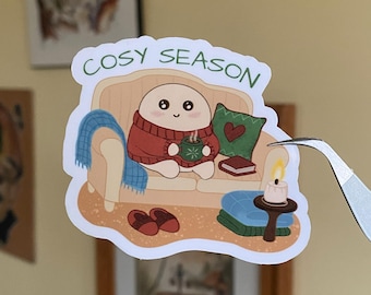 May include: A sticker featuring a cartoon character in a red sweater, seated on a beige couch, holding a mug. The sticker has the text "COSY SEASON" and includes a candle, books, and a blanket. The sticker is being held by tweezers.
