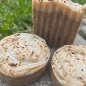 May include: Three handmade soaps. Two round soaps and one rectangular bar soap. The soaps are light brown with a swirl design on top and dark brown speckles. The soaps are on a gray stone surface with green grass in the background.