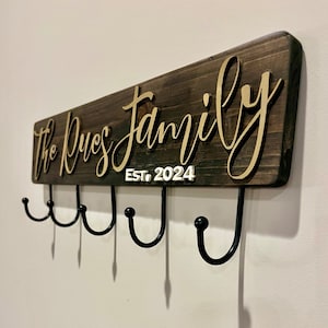 May include: A wooden sign with the text "The Dues Family Est. 2024" in gold lettering. The sign has five black metal hooks attached to the bottom.
