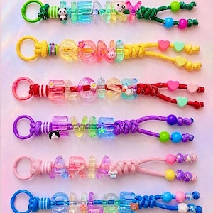 May include: Six colourful keychains featuring plastic alphabet beads spelling the names HENRY, QONI, ELOISE, EVIE, ARIA, and CHLOE. Each keychain includes a plastic ring, various translucent letter beads, small animal or flower charms, and knotted cord ends with round accent beads in matching colour palettes.