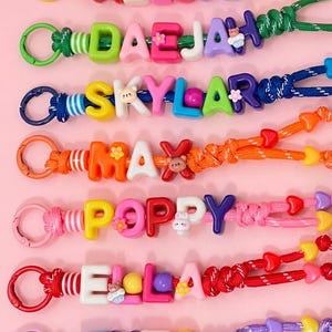 May include: Seven colourful keychains featuring plastic alphabet beads spelling the names ABIGAIL, DAEJAH, SKYLAR, MAX, POPPY, ELLA, and THEO. Each keychain includes a plastic clasp, small decorative charms, and a braided nylon cord with heart beads.