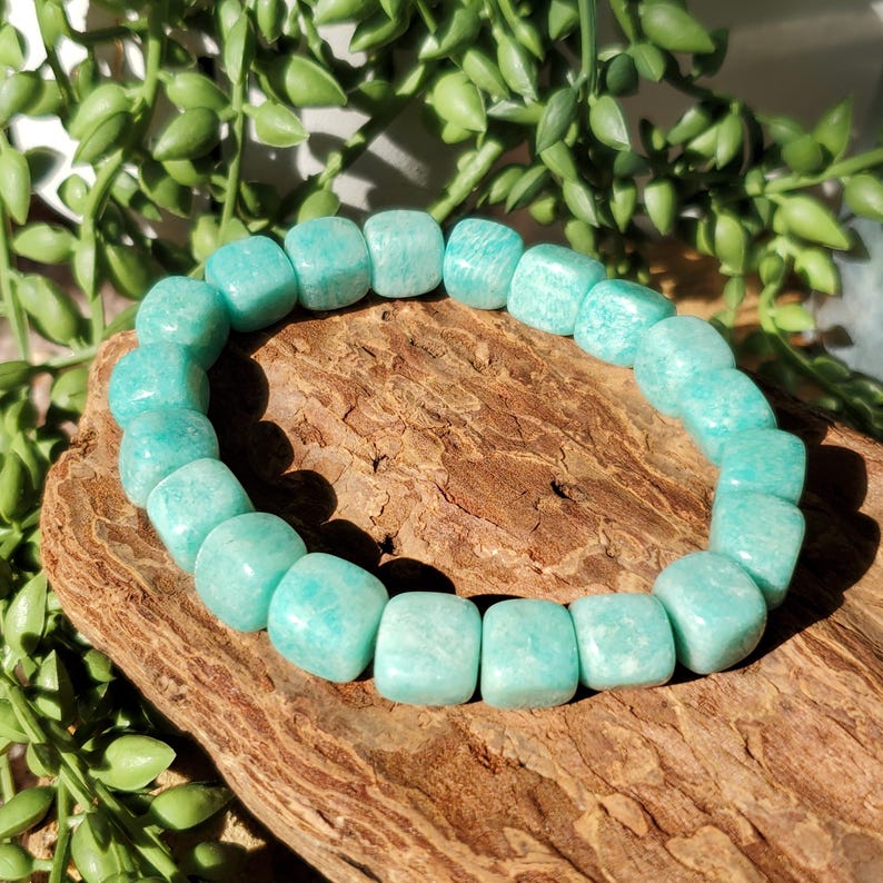 Puede incluir: Un brazalete de amazonita turquesa. El brazalete est&aacute; hecho de cuentas cuadradas, ensartadas para formar una forma circular. Las cuentas son de color azul verdoso claro. El brazalete descansa sobre un trozo de madera, con follaje verde en el fondo.