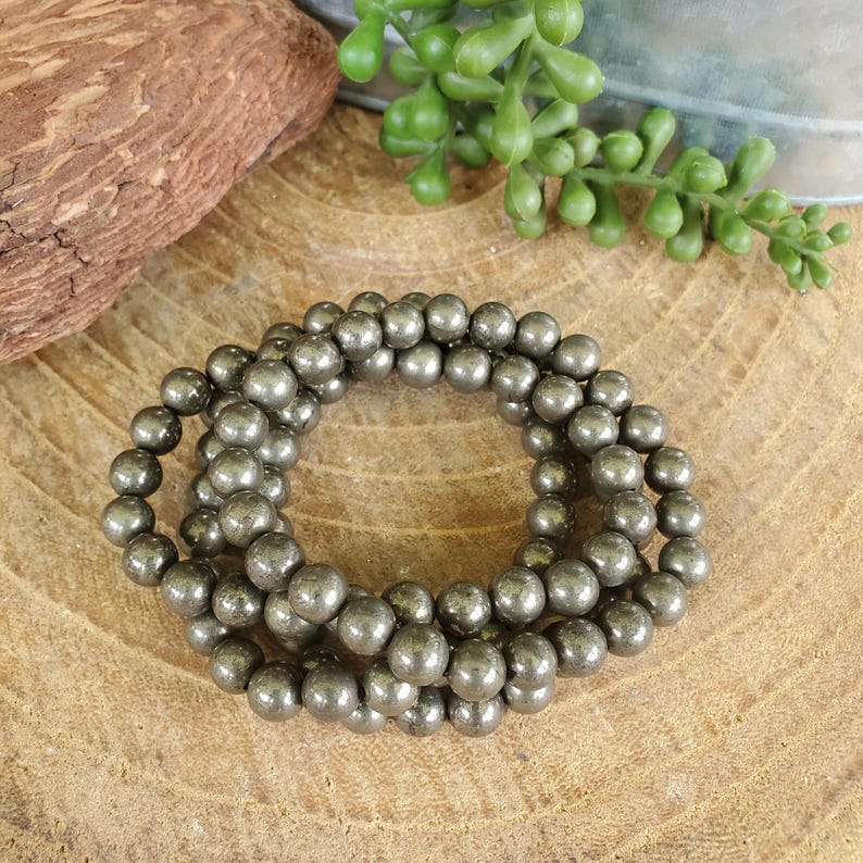 May include: A set of three pyrite bead bracelets. The beads are a metallic gray color and are strung together. The bracelets are displayed on a wooden surface with a piece of wood and greenery in the background.