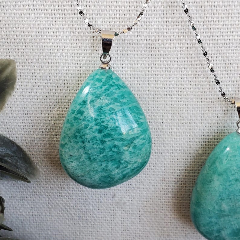 May include: Close-up of two necklaces featuring teardrop pendants. The pendants are a marbled turquoise hue, suspended from silver chains. The necklaces are displayed against a white, textured backdrop. These are examples of jewellery.