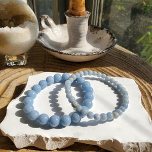 May include: Two light blue beaded bracelets rest on a white, textured surface. The bracelets are made of round, polished beads. A candle holder with a lit candle and a geode are in the background, suggesting a spiritual or decorative theme.