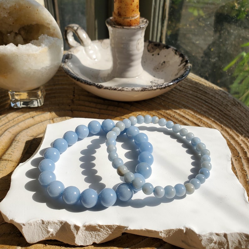 May include: Two light blue beaded bracelets rest on a white, textured surface. The bracelets are made of round, polished beads. A candle holder with a lit candle and a geode are in the background, suggesting a spiritual or decorative theme.