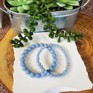May include: Two light blue beaded bracelets of varying sizes rest on a white surface. The bracelets are made of round, polished beads. The background includes a small metal bucket with green plants and a wooden slice.