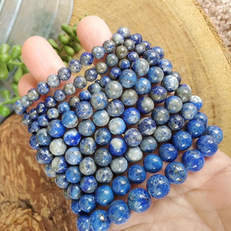 May include: Several strands of blue lapis lazuli gemstone beads in varying sizes. The beads have a mix of deep blue and gray tones, with a polished, round shape. The beads are held in a hand.