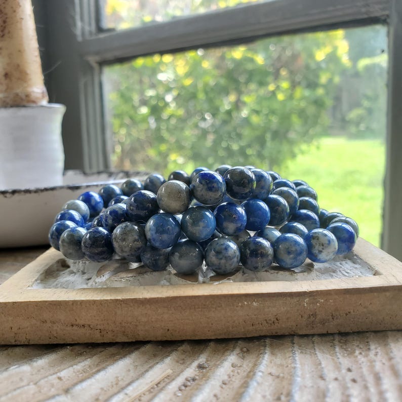 May include: A stack of blue and gray beaded bracelets rests on a light brown, square tray. The beads are round and have a marbled appearance. The background includes a window with a view of green foliage.