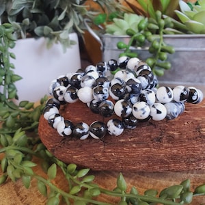 May include: Three beaded bracelets with black and white patterned beads are displayed on a piece of wood. The bracelets are made of round beads with a marbled design. Green plants and a white pot are in the background.