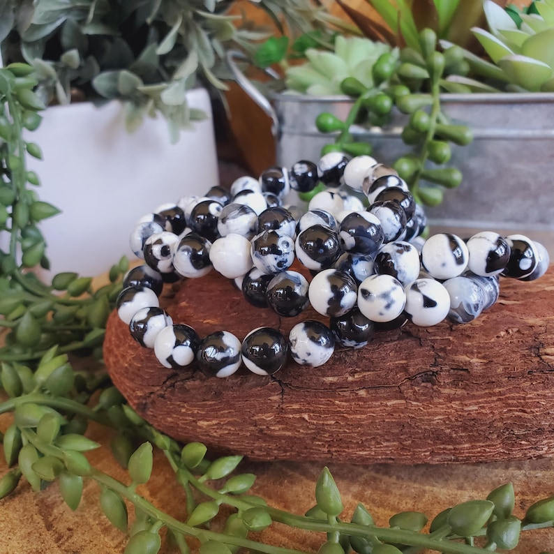 May include: Three beaded bracelets with black and white patterned beads are displayed on a piece of wood. The bracelets are made of round beads with a marbled design. Green plants and a white pot are in the background.