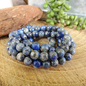 May include: A collection of round, blue and gray gemstone beads. The beads are a mix of solid blue and speckled gray, arranged in a circular pattern. The beads are likely used for jewelry making.