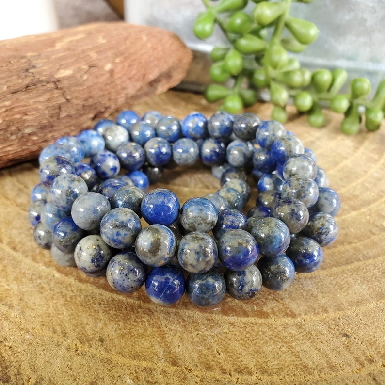 May include: A collection of round, blue and gray gemstone beads. The beads are a mix of solid blue and speckled gray, arranged in a circular pattern. The beads are likely used for jewelry making.