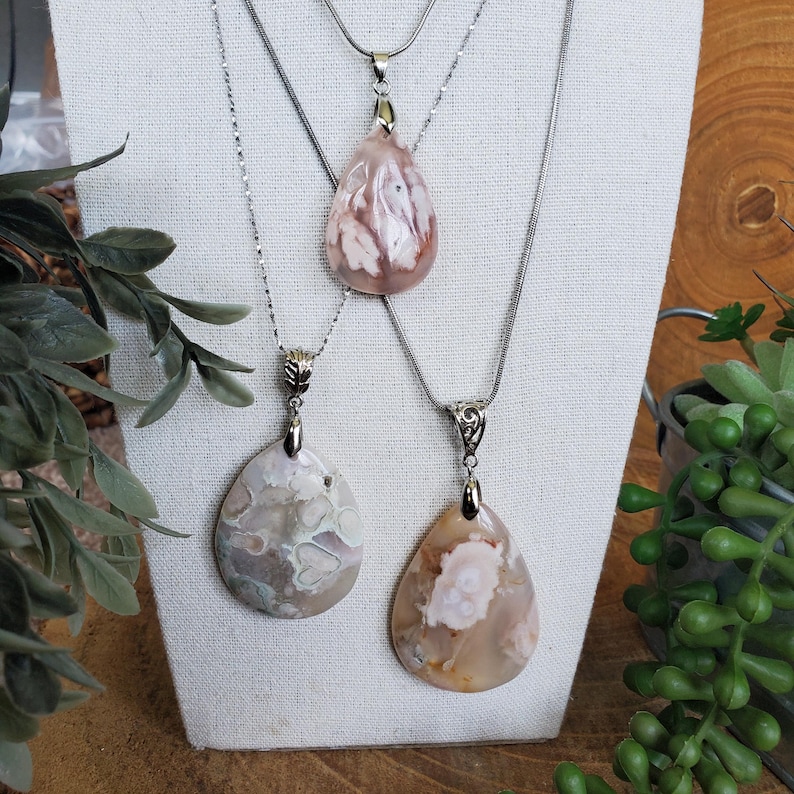 May include: Three gemstone pendant necklaces displayed on a white fabric. The pendants are teardrop-shaped, with silver-colored chains. The gemstones have a mix of pink, white, and brown colors. The necklaces are displayed on a white fabric.