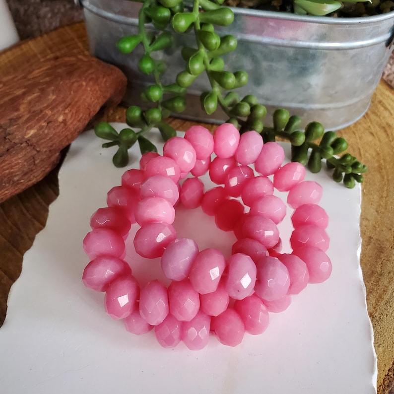 May include: A close-up of three strands of pink faceted beads. The beads are a light, translucent pink color and are arranged in a circular pattern. The background includes greenery and a metal container.