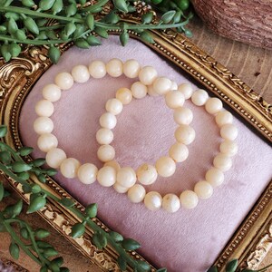May include: Two cream-colored beaded bracelets rest on a pink velvet surface within a gold-colored ornate frame. Green leafy vines are arranged around the frame. The bracelets are made of round, smooth beads.