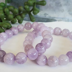 K&ouml;nnte beinhalten: Zwei Armb&auml;nder mit runden, polierten Lavendel-Amethyst-Edelsteinen. Die Armb&auml;nder sind aufgef&auml;delt und zeigen die nat&uuml;rlichen Farbvariationen der Steine. Der Hintergrund umfasst eine gr&uuml;ne Pflanze und eine wei&szlig;e Oberfl&auml;che.