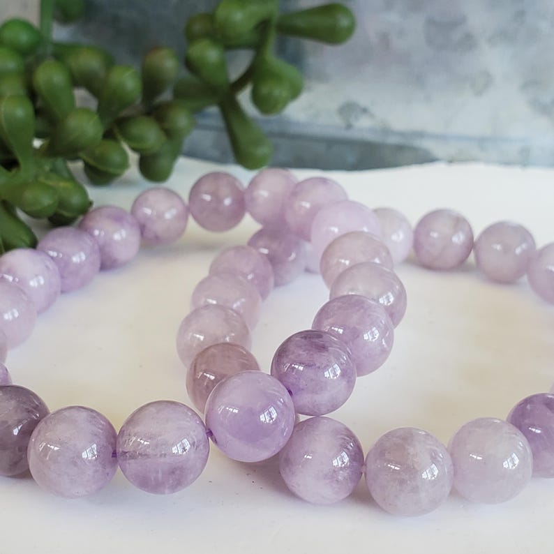 K&ouml;nnte beinhalten: Zwei Armb&auml;nder mit runden, polierten Lavendel-Amethyst-Edelsteinen. Die Armb&auml;nder sind aufgef&auml;delt und zeigen die nat&uuml;rlichen Farbvariationen der Steine. Der Hintergrund umfasst eine gr&uuml;ne Pflanze und eine wei&szlig;e Oberfl&auml;che.