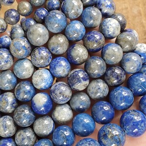 May include: A collection of round, polished lapis lazuli beads in varying shades of blue and gray. The beads have a speckled appearance with natural variations in color and texture, perfect for jewelry making.