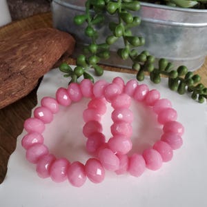 May include: Two pink beaded bracelets with faceted beads. The bracelets are displayed on a white surface, with a wooden piece and a metal container with green plant stems in the background. The bracelets are a light, bright pink color.