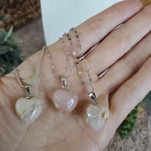 Op de afbeelding: Drie hartvormige hangerkettingen. De hangers zijn gemaakt van gepolijste steen in tinten wit, roze en cr&egrave;me. Elke hanger hangt aan een zilverkleurige ketting. De kettingen worden op een hand getoond.