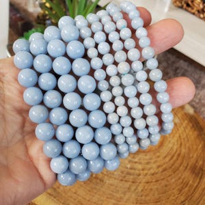 May include: Several strands of light blue gemstone beads, varying in size, are held in a hand. The beads have a smooth, polished surface and a consistent, pale blue color. The bracelets are likely for jewelry making or wearing.