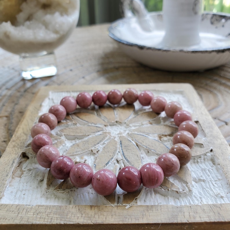 May include: A pink gemstone bracelet with round beads. The bracelet is on a wooden surface with a carved floral design.