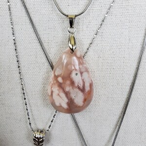 May include: A teardrop-shaped pendant necklace with a silver chain. The pendant is a light pink stone with white and brown patterns. The necklace is displayed on a white surface.