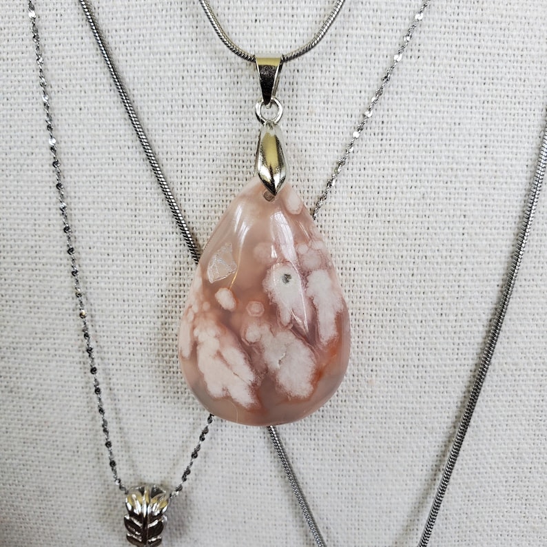 May include: A teardrop-shaped pendant necklace with a silver chain. The pendant is a light pink stone with white and brown patterns. The necklace is displayed on a white surface.