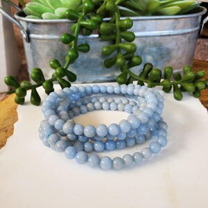 May include: Several light blue beaded bracelets are coiled on a white surface. The bracelets are made of small, round beads. Green leafy plants and a metal container are in the background.