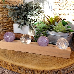 May include: Four crystal spheres, two amethyst and two clear quartz, are displayed on a light wood stand. The spheres are arranged in a row, with greenery and a woven basket in the background. The spheres are approximately 2 inches in diameter.